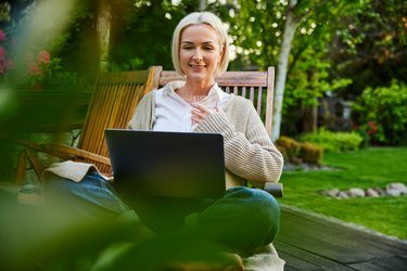 Adult woman sitting relaxed with laptop on garden terrace during summertime